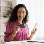 Una mujer sonriente sostiene una tarjeta de crédito en una mano y un teléfono móvil en la otra, mientras disfruta de su experiencia de compra en línea.
