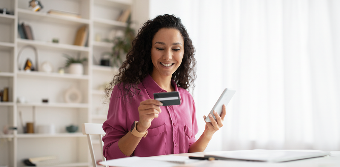 Una mujer sonriente sostiene una tarjeta de crédito en una mano y un teléfono móvil en la otra, mientras disfruta de su experiencia de compra en línea.